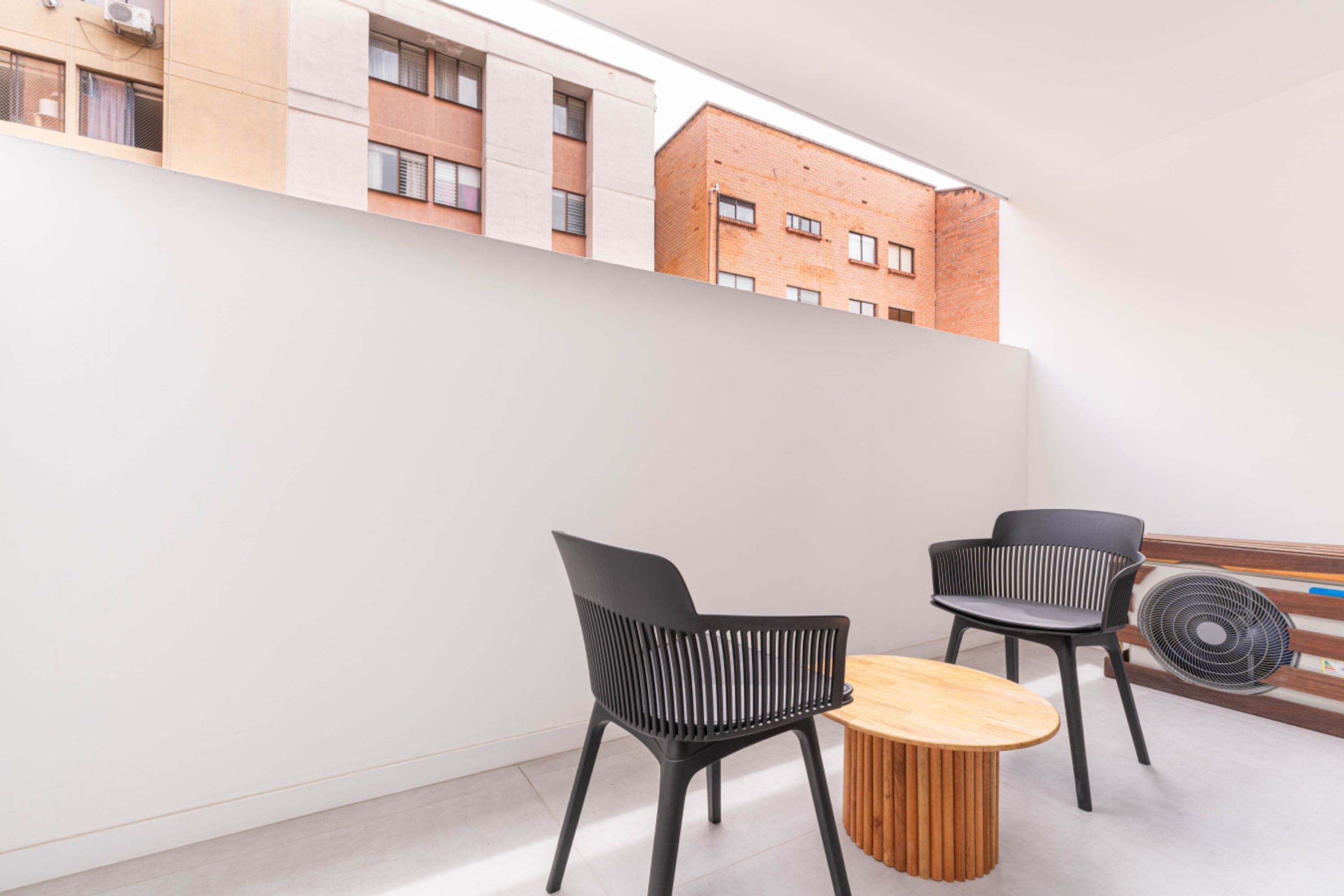 Ein moderner Balkon mit zwei schwarzen Stühlen und einem Holztisch, mit Blick auf eine Stadtlandschaft mit Backsteingebäuden.