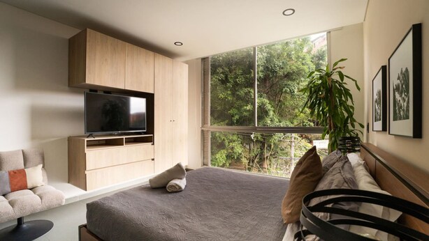 Un dormitorio moderno con una cama grande, un televisor montado en la pared, un sofá y una ventana con vistas a árboles.