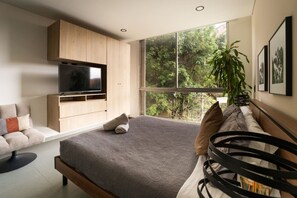 Une chambre moderne avec un grand lit, un téléviseur fixé au mur, un canapé et une fenêtre donnant sur les arbres.