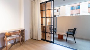 Ein modernes Zimmer mit einer Holzlounge, einem runden Tisch und einer Glastür, die zu einem Außenbereich mit Stühlen und einem Gebäude im Hintergrund führt.