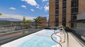 A rooftop pool with clear blue water, surrounded by glass railings, and a view of a tall building and mountains in the distance. - Metropolitan Living - 1615 (Medellín)