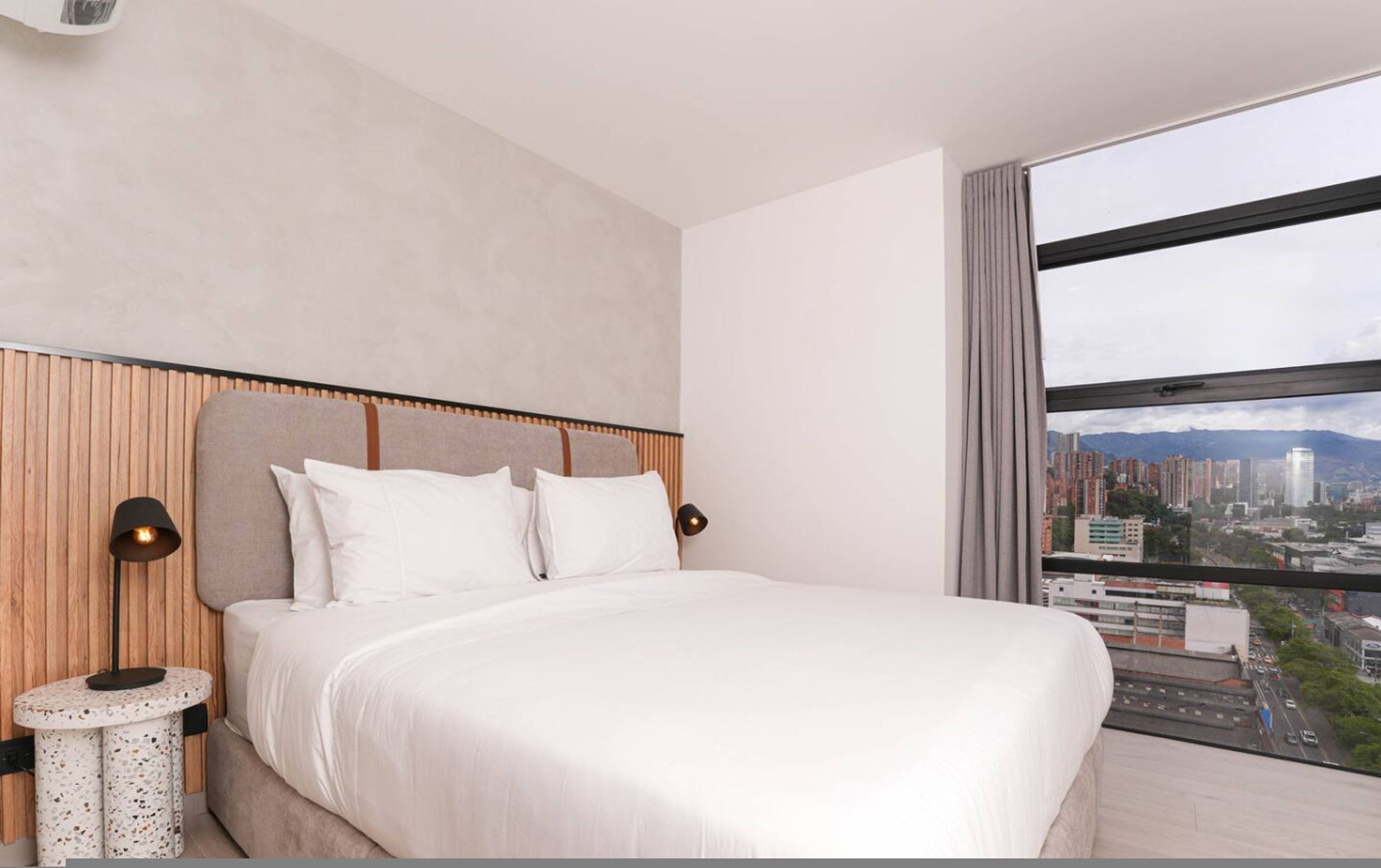 A modern hotel room with a large bed, a city view, and a wooden headboard.