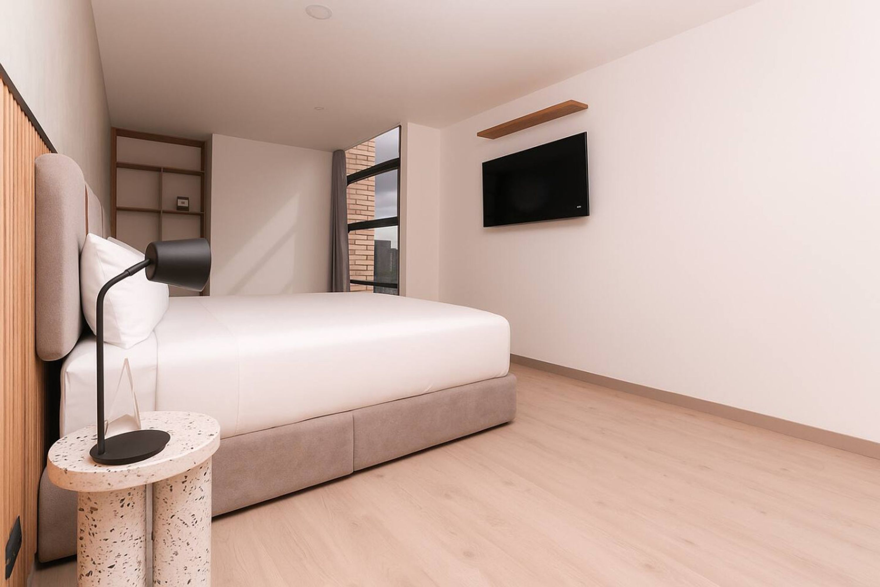 Un dormitorio moderno con una cama, una mesita redonda, un televisor montado en la pared y un ventanal con vistas a edificios.