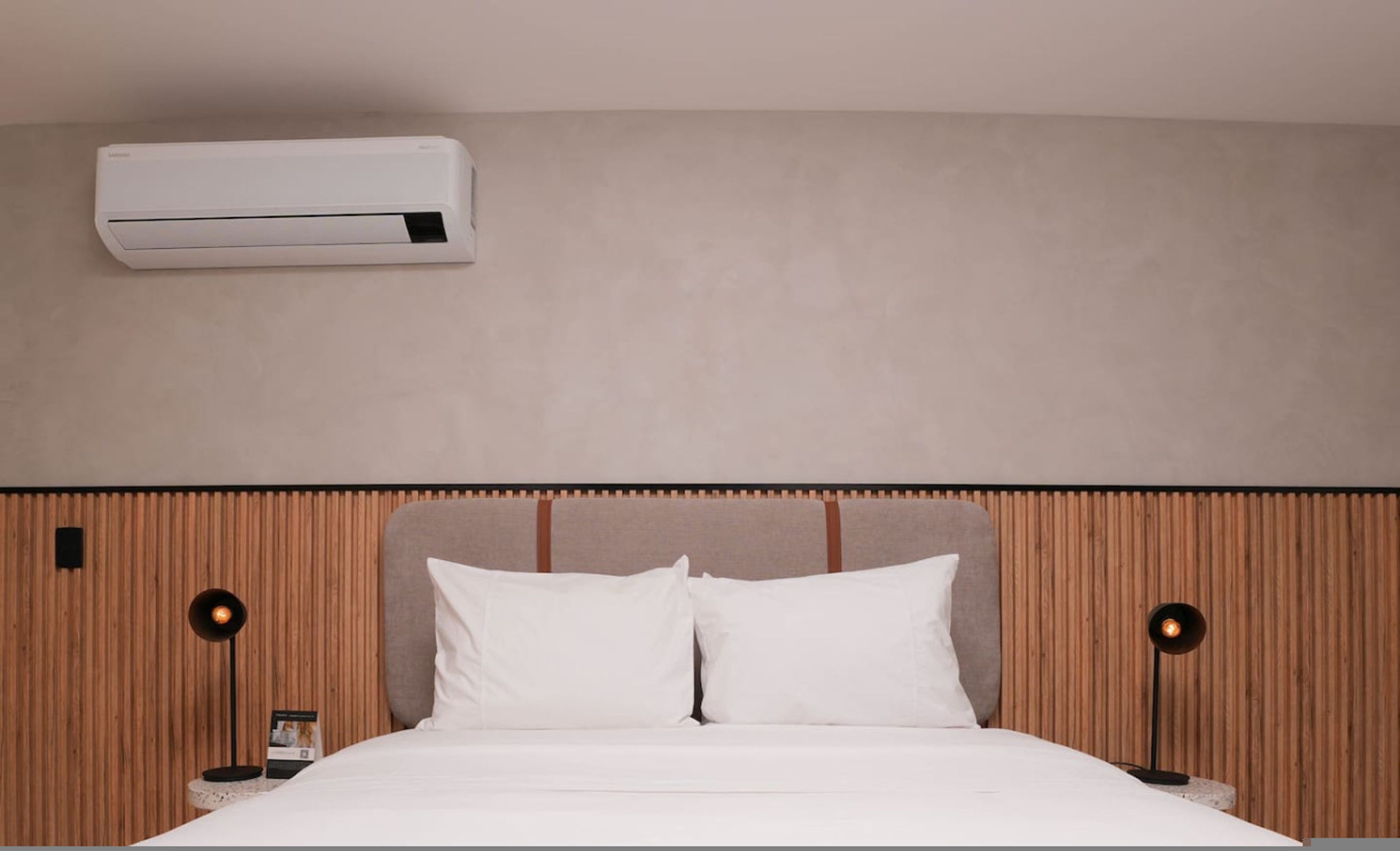 A hotel room with a bed, white bedding, a wooden headboard, two bedside lamps, and a wall-mounted air conditioner.