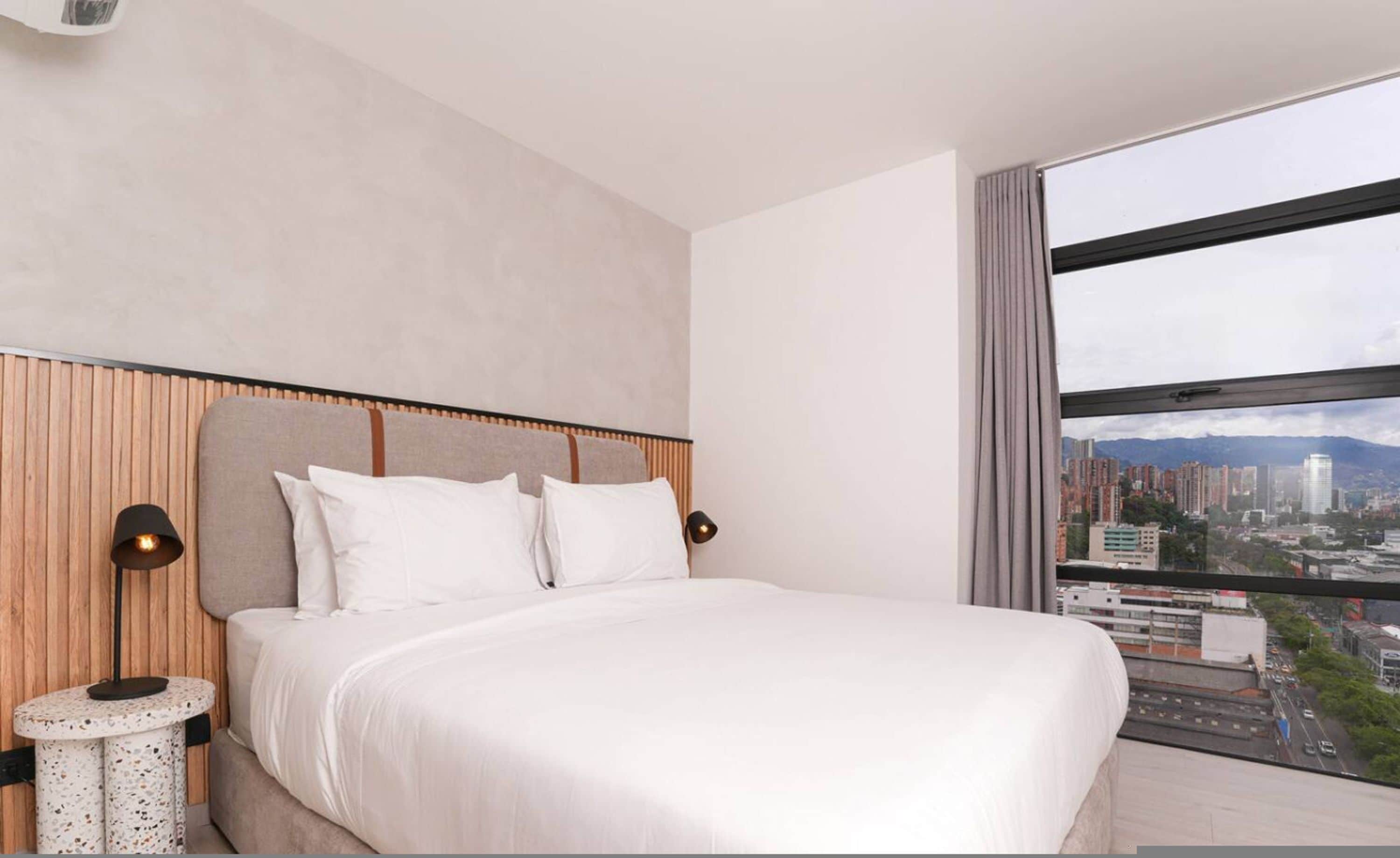 A modern hotel room with a large bed, two bedside lamps, a round side table, and a city view through a large window.