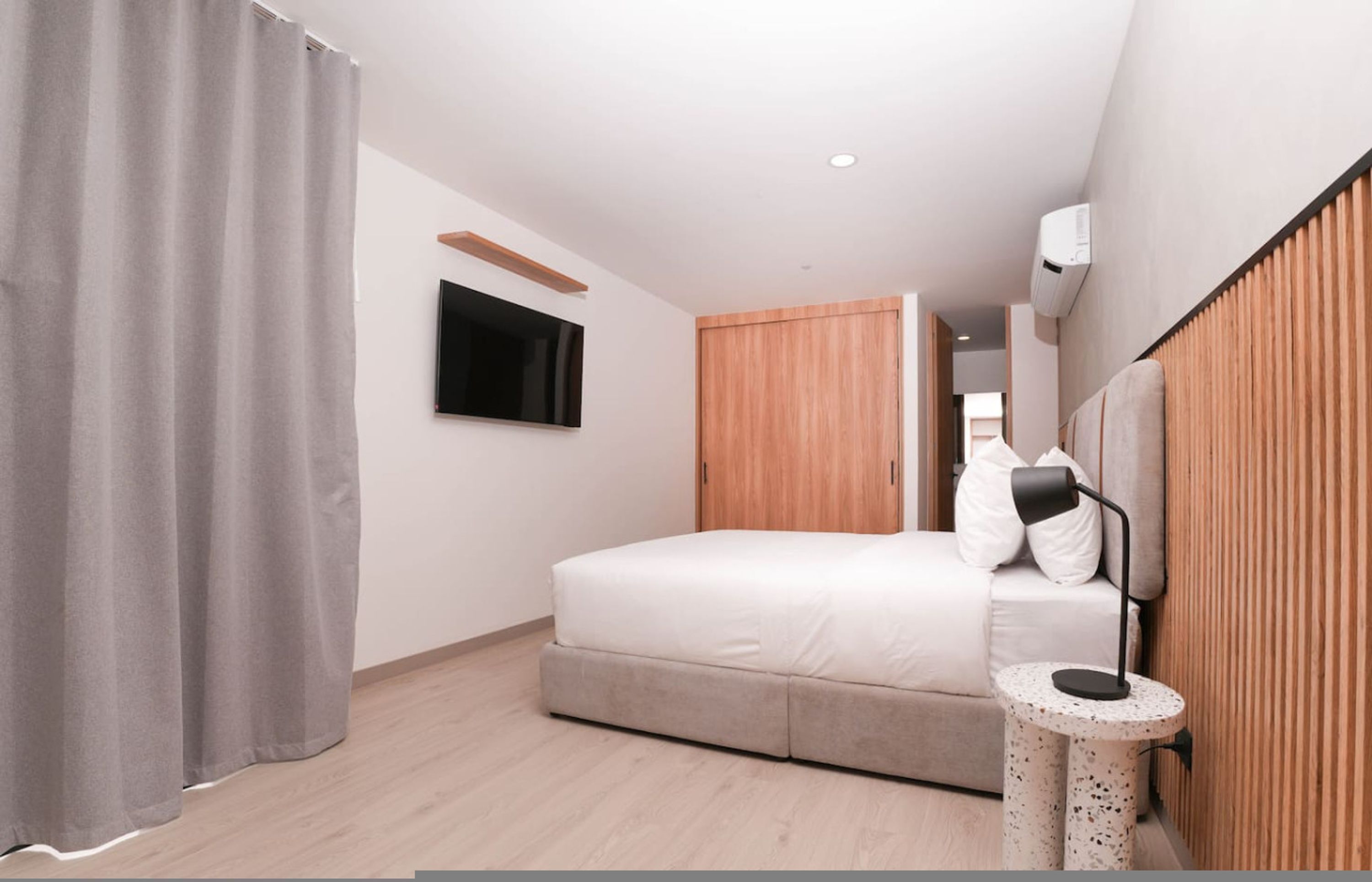 Een moderne slaapkamer met een bed, een aan de muur bevestigde televisie, een houten garderobe en een nachtkastje met een lamp.