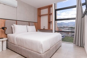 Un dormitorio moderno con una cama grande, una mesita de noche, un estante empotrado y vistas a un paisaje urbano a través de un ventanal.
