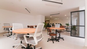Una sala de conferencias moderna con mesas redondas, sillas blancas y marrones, y plantas en macetas.