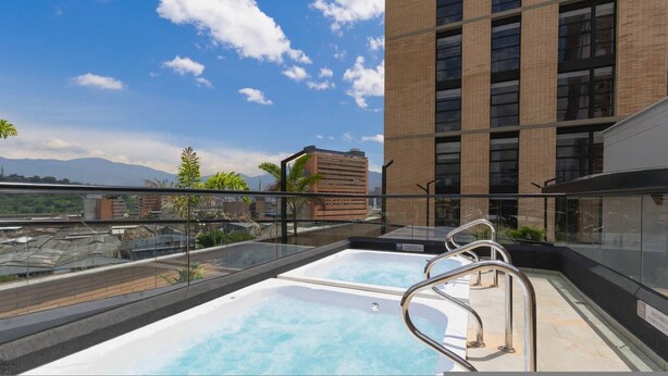 Una piscina en la azotea con barandillas de metal, cielo despejado y vistas de un paisaje urbano con montañas al fondo.