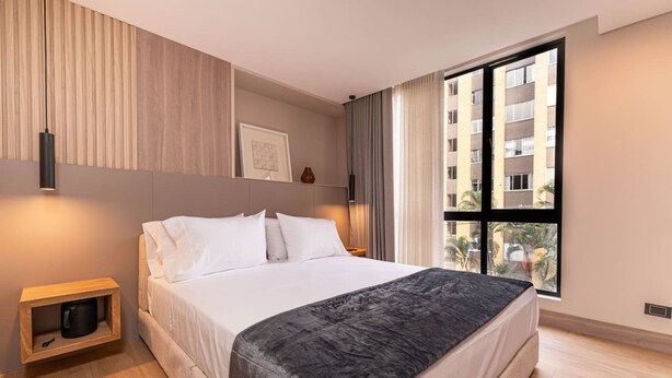 A modern hotel room with a large bed, a bedside table, a wall-mounted light, a window with curtains, and a view of buildings outside. - Jatisa 303 in Medell n (Medellín)
