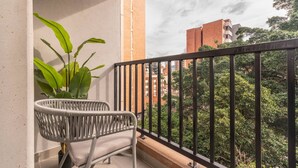 Un balcón con una silla de mimbre blanca, una planta en maceta y vistas a edificios y árboles.