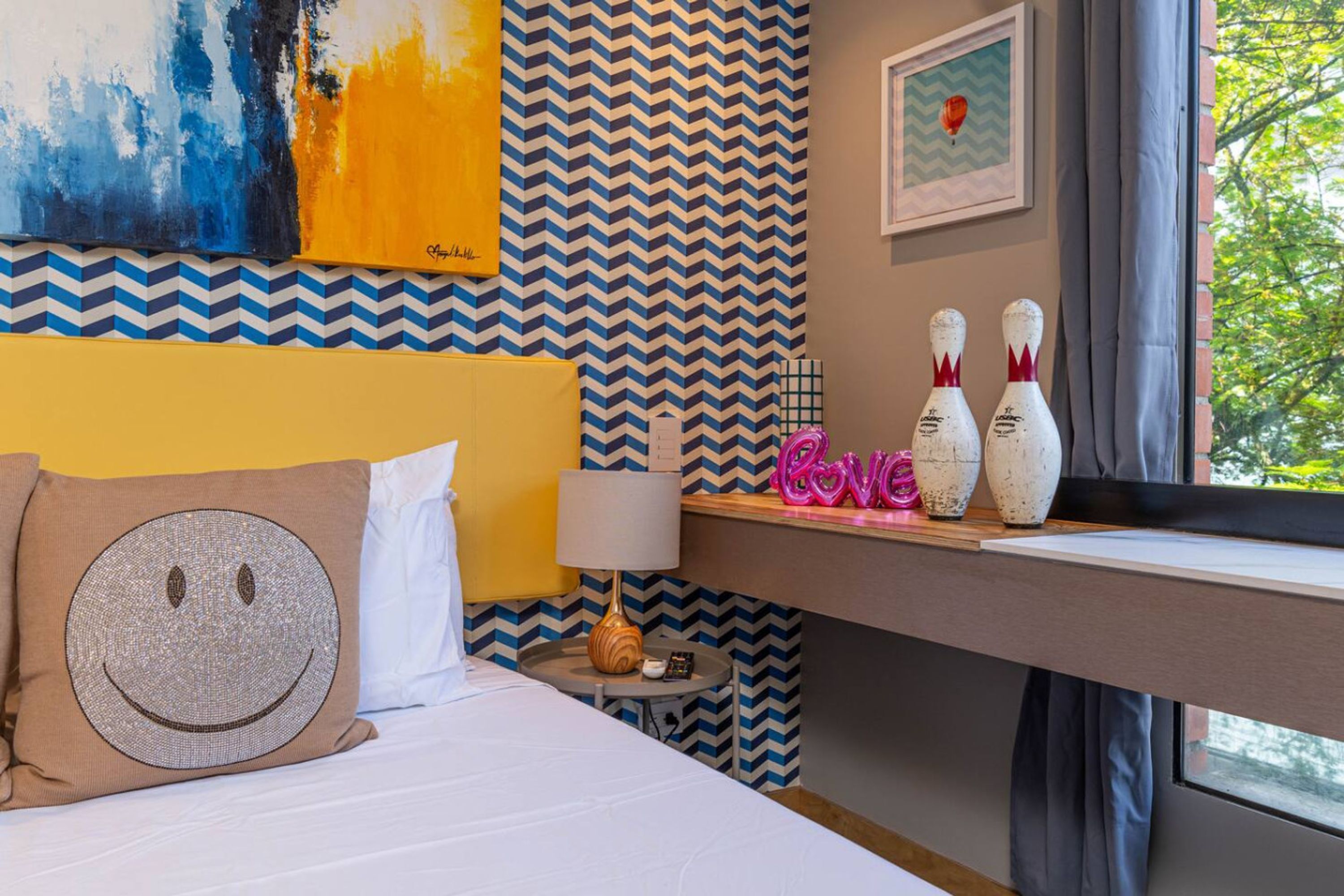 Une chambre d’hôtel avec un lit, une table de chevet avec une lampe, un coussin décoratif, un mur orné d’un motif en chevrons et une fenêtre donnant sur un paysage.