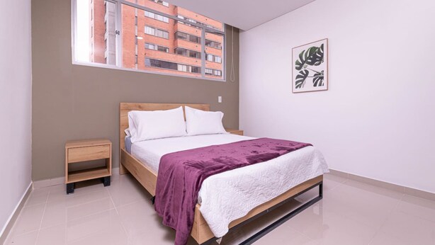 Une chambre à coucher avec un lit, une table de chevet, une fenêtre donnant sur des immeubles et un tableau encadré au mur.