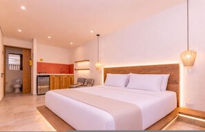 Un dormitorio con una cama grande, cabecera de madera, mesita de noche, una silla, una pequeña kitchenette y un baño.