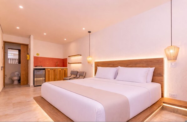 Habitación de hotel con una cama grande, cabecera de madera, una kitchenette pequeña y un baño visible a través de una puerta abierta.
