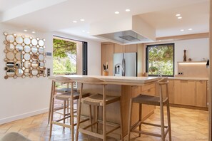 A modern kitchen with a central island, bar stools, and a decorative wall piece. - VILLA MAROMA By Terry's Homes (Saint-Tropez)