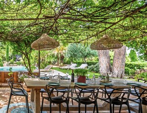 An outdoor dining area with wicker umbrellas, a pool, and lounge chairs in a lush garden setting. - VILLA MAROMA By Terry's Homes (Saint-Tropez)