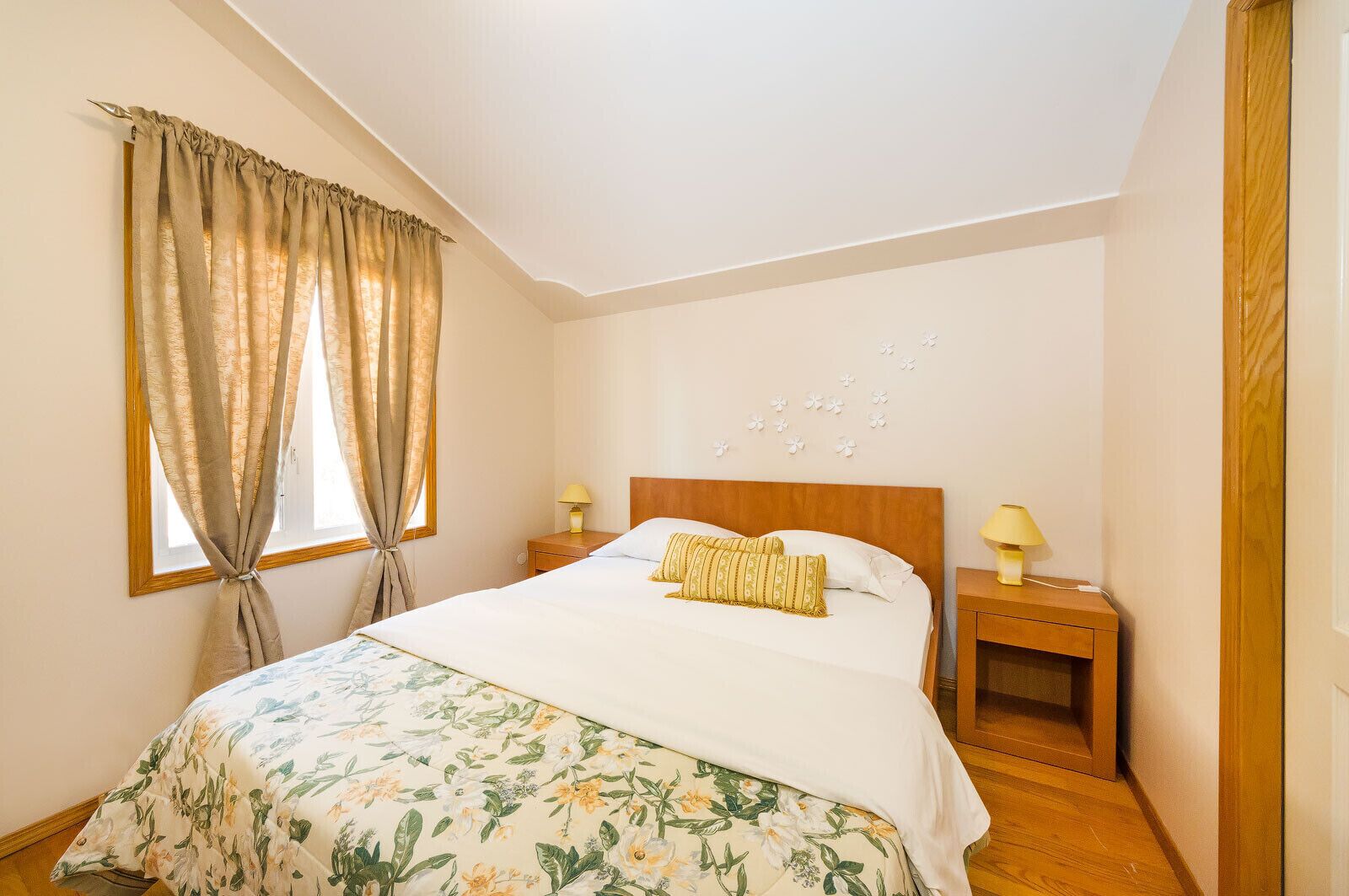 Un dormitorio con una cama, dos mesitas de noche, una ventana con cortinas y una lámpara fijada a la pared.