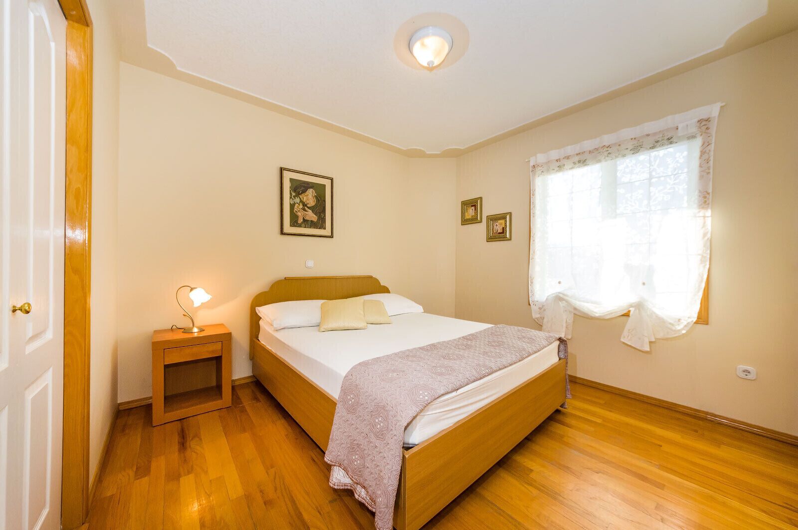 A bedroom with a wooden bed, a nightstand with a lamp, a window with curtains, and a painting on the wall.