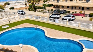 Una piscina de diseño curvo, rodeada por una valla blanca, con vista a coches estacionados y edificios al fondo.