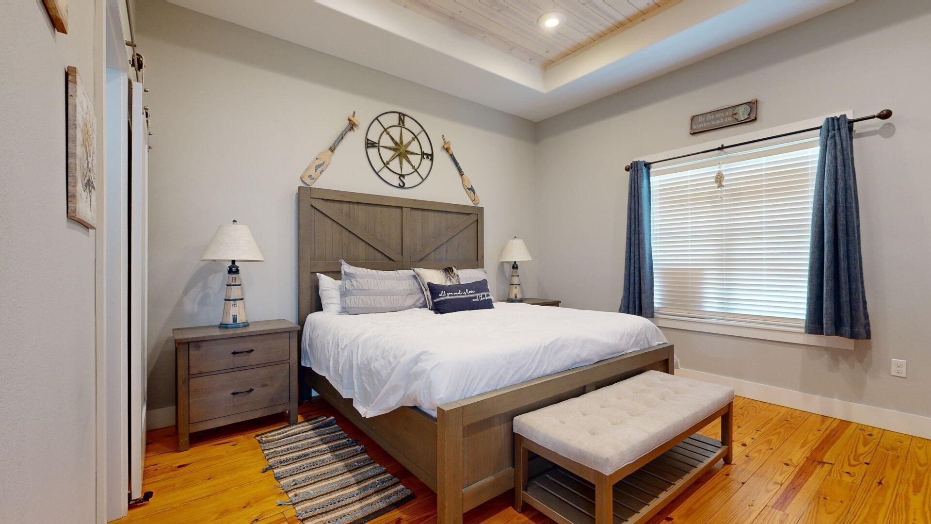 Un dormitorio con cama de madera, mesitas de noche, un banco, una ventana con cortinas y un techo con vigas de madera.