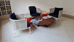 A room with a round wooden table, two white wicker chairs with blue cushions, and a small orange stool. - Jardins do Atlântico - Miramar  (João Pessoa)