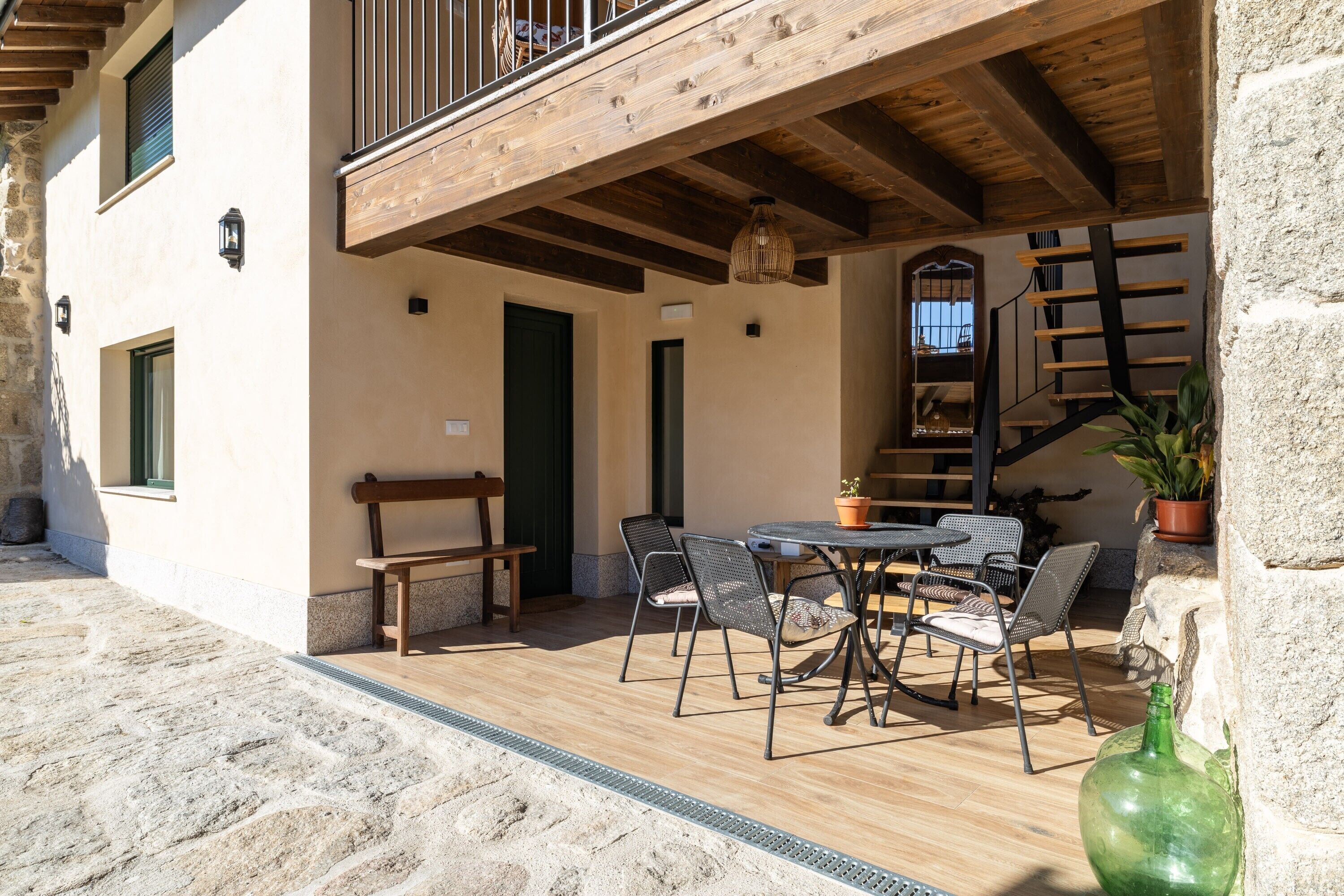 Eine Terrasse im Freien mit einem Holztisch und -stühlen, einer Bank, einem Blumentopf und einer Treppe, die zu einer oberen Ebene führt.