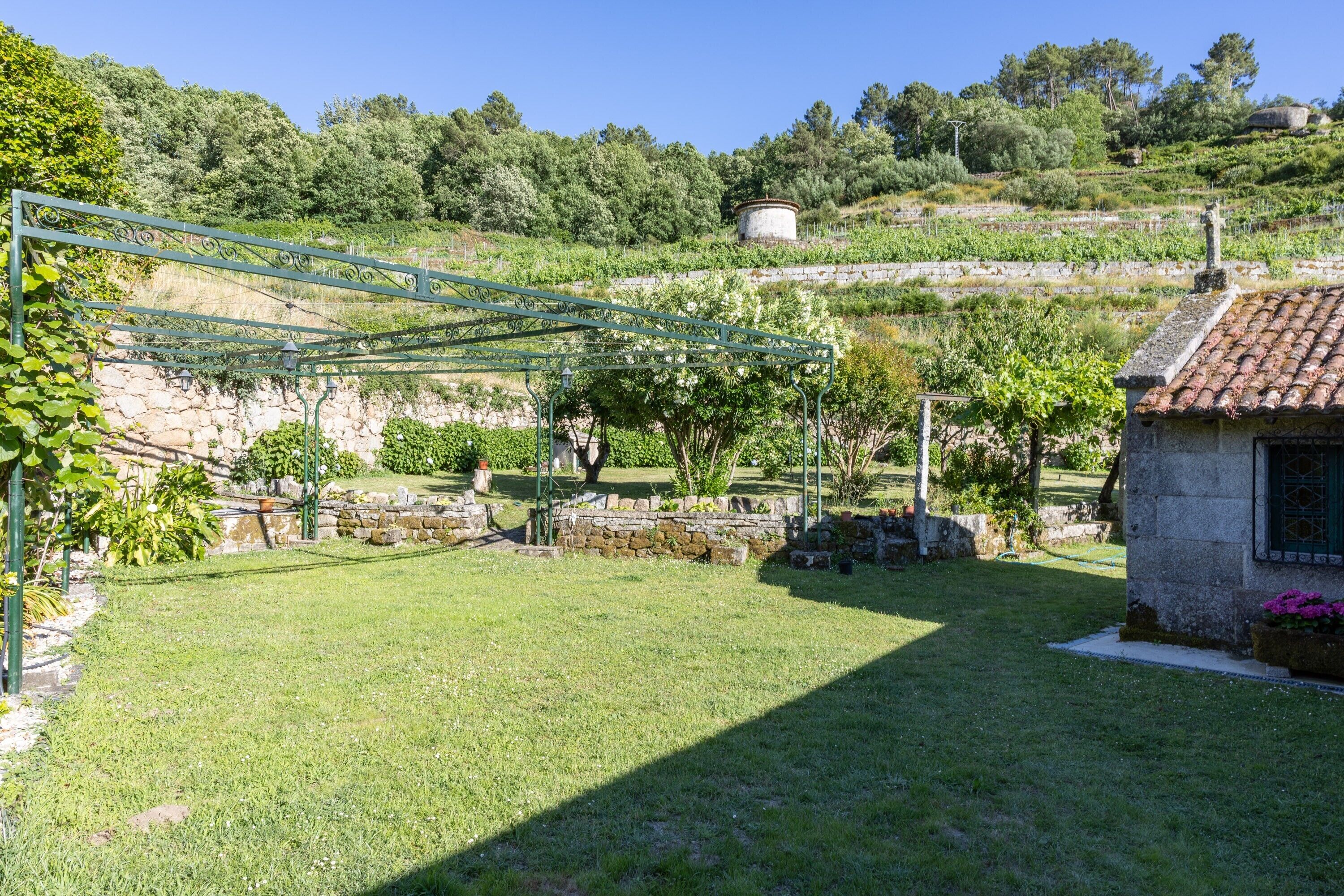 A garden with a stone wall, a pergola, and a small building with a tiled roof.