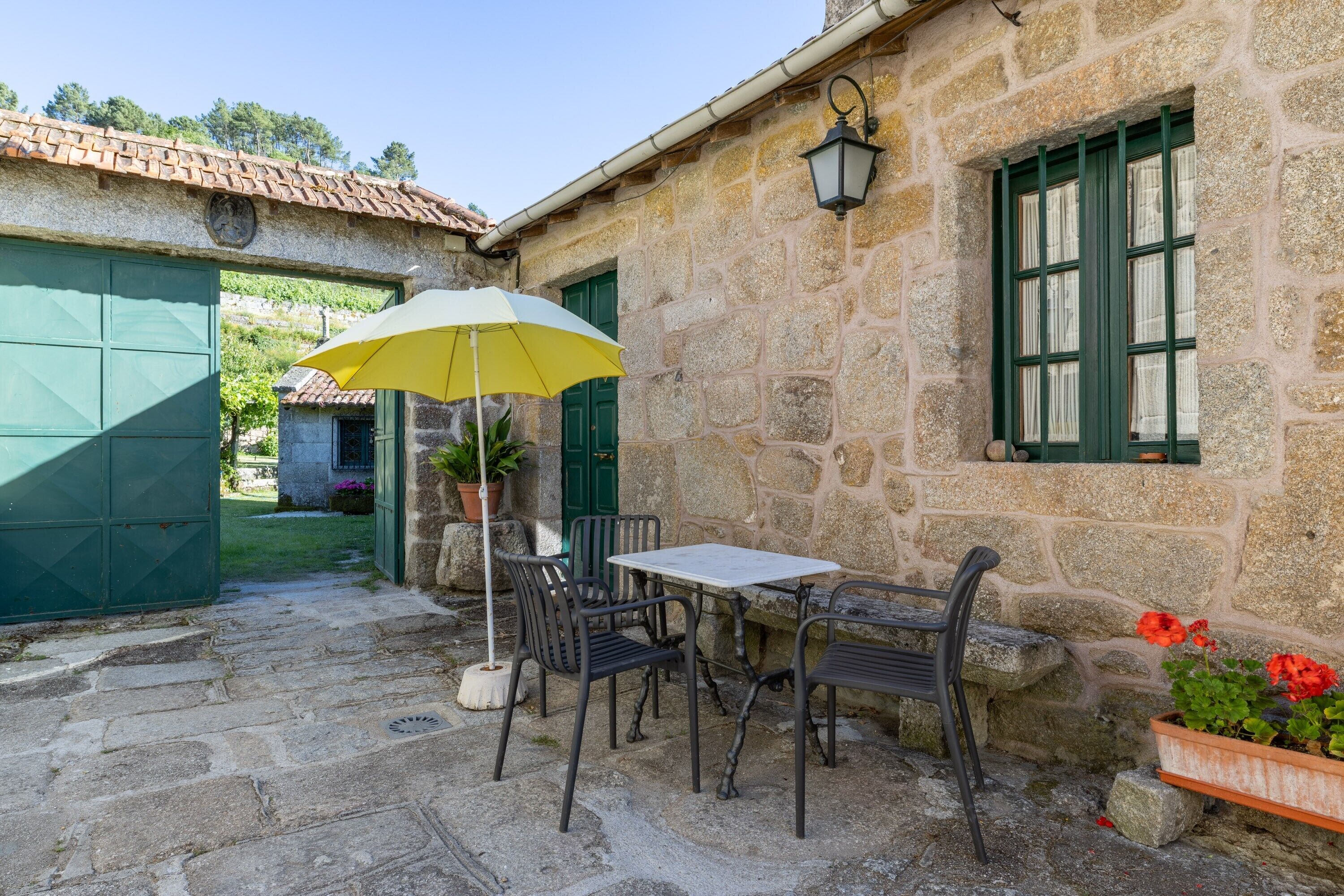 A stone building with a tiled roof, a table with chairs, a yellow umbrella, and a potted plant.