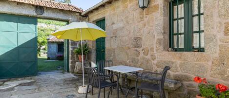 A stone building with a tiled roof, a table with chairs, a yellow umbrella, and a potted plant.