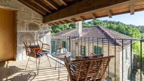 Un balcón con muros de piedra, vigas de madera, muebles de mimbre y vistas a un edificio con tejado de tejas rojas.