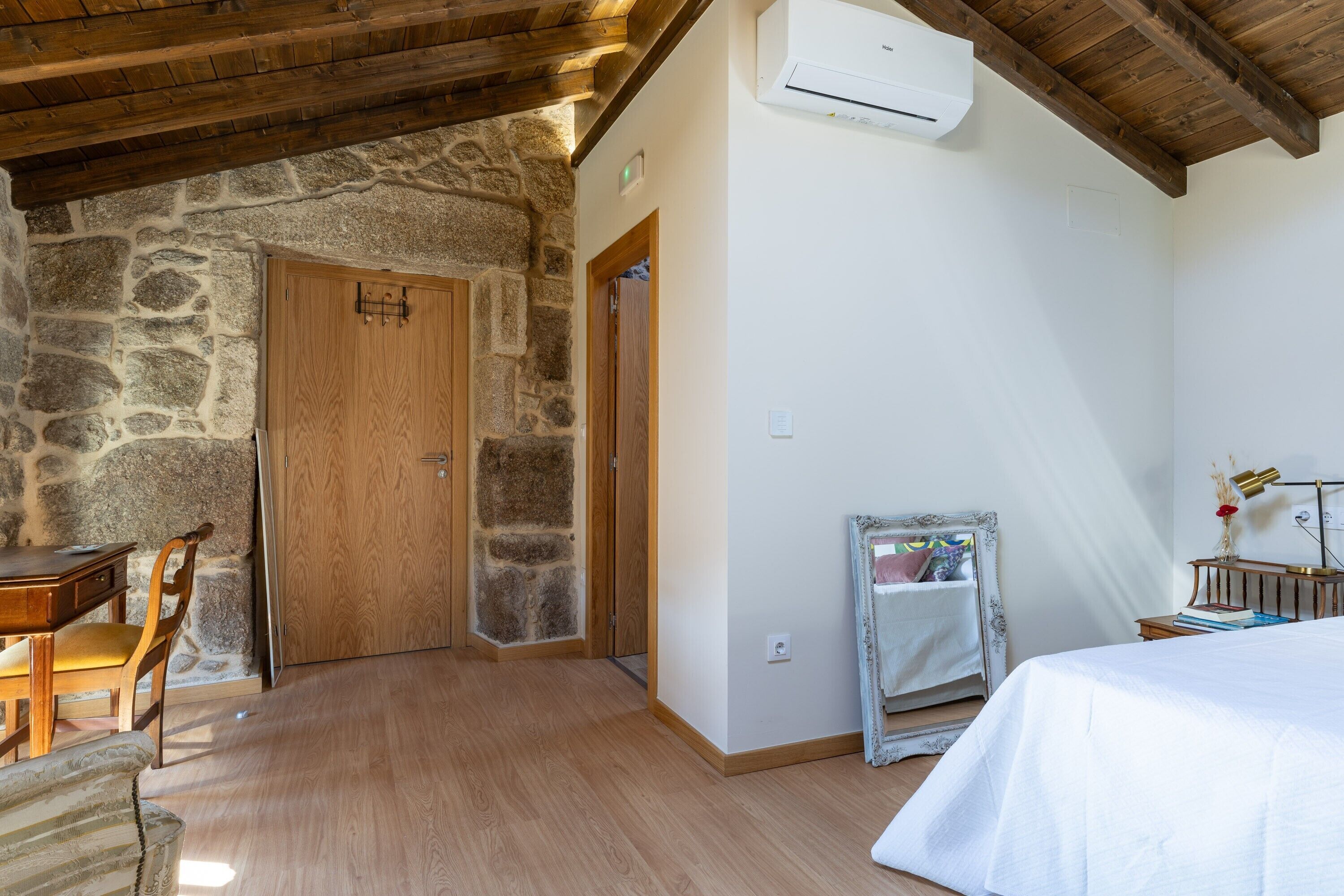 Une pièce avec un mur en pierre, une porte en bois, un lit, une table de chevet, une lampe et un miroir.