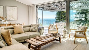 Ein modernes Wohnzimmer mit Sofa, Couchtisch und Sesseln, ein Essbereich mit Tischen und Stühlen sowie eine große Glasschiebetür, die auf einen Balkon mit Meerblick führt.