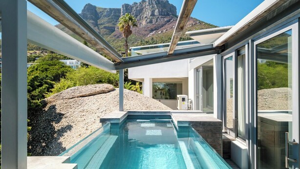 Ein moderner Außenpool mit klarem, blauem Wasser, umgeben von einem weißen Gebäude mit großen Glasfenstern, vor einer Bergkulisse.