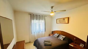 Ein Schlafzimmer mit Bett, Nachttisch, Deckenventilator, Fenster mit Vorhängen und einem gerahmten Bild an der Wand.