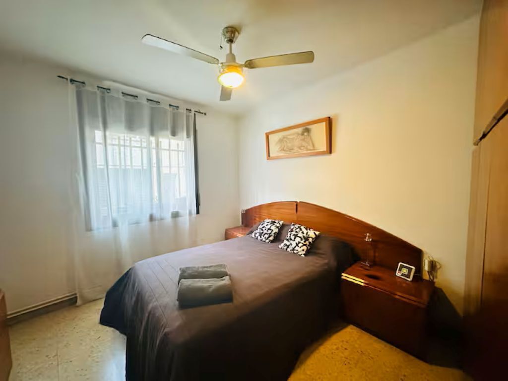 Quarto com cama, mesa de cabeceira, ventoinha no teto, janela com cortinas e quadro na parede.