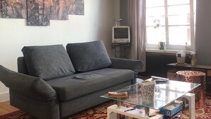 A living room with a grey sofa, a glass coffee table, a cityscape painting, a television, and a large window with curtains. - Godewind 1 Apartments with Wi-Fi (Jever)