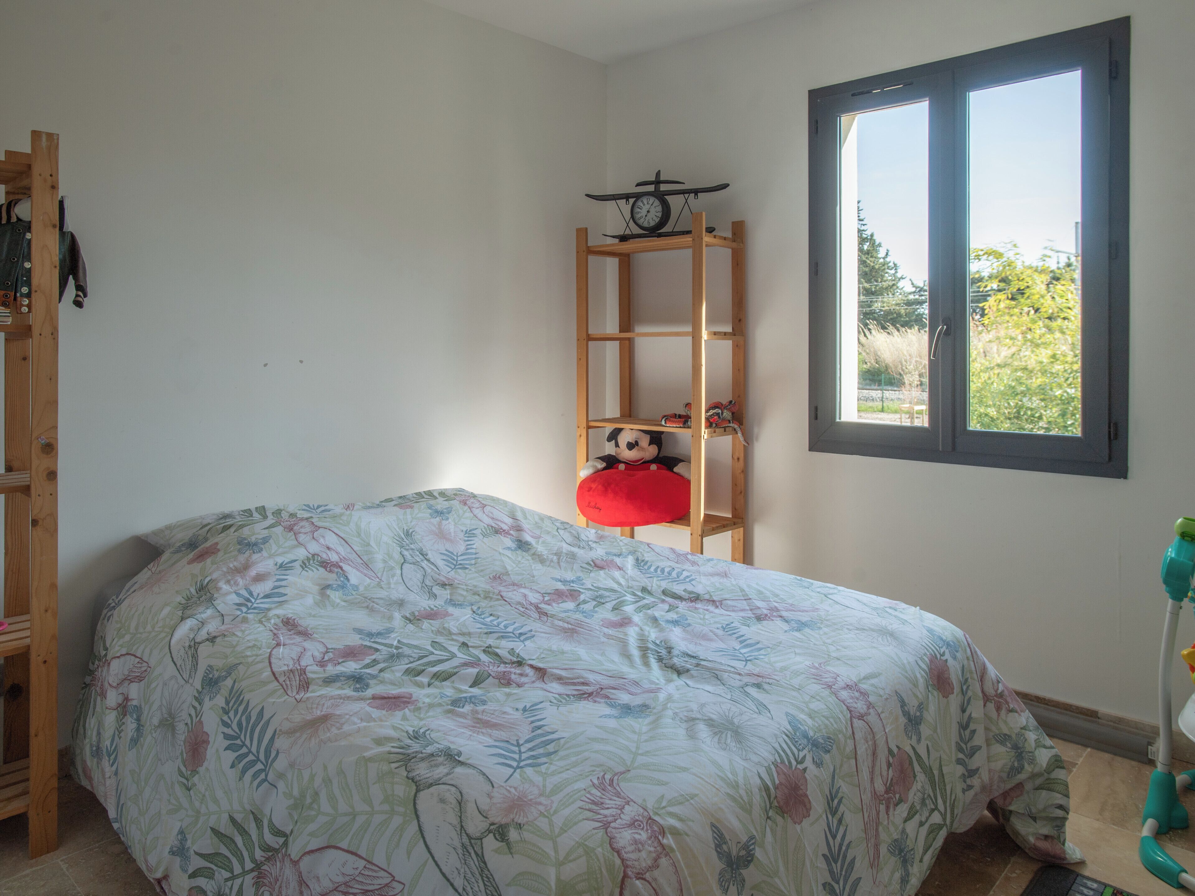 A bedroom with a floral bedspread, a wooden ladder shelf, a Mickey Mouse plush toy, a clock, and a window with a view of greenery.