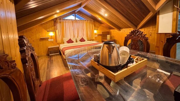 A cozy attic bedroom with a wooden ceiling, a bed with red bedding, a glass table with a kettle, and a window with curtains. - Hotel The Red Apple  (Manali)