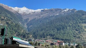 A mountainous landscape with snow-capped peaks, a dense forest, and a small settlement with buildings. - Hotel The Red Apple  (Manali)