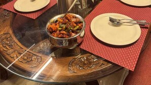 A dining table with a glass top, surrounded by wooden chairs, set with plates, cutlery, and a bowl of food. - Hotel The Red Apple  (Manali)