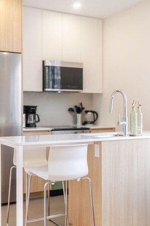 A modern kitchen with a stainless steel refrigerator, a microwave, a sink with a faucet, and two bar stools. - Chic Urban Escape – 2BR Retreat Montreal Living (Montréal)