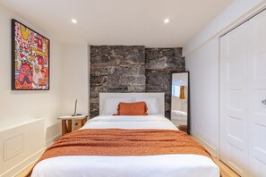 A bedroom with a stone wall, a bed with white bedding, a brown throw blanket, a nightstand, a mirror, and a colorful artwork on the wall. - Grand Urban Retreat – Spacious 5BR Heart MTL (Montréal)