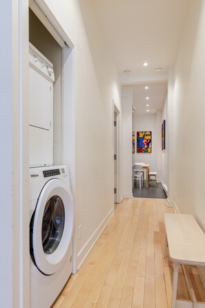 A laundry room with a front-loading washer and dryer, a wooden floor, and a dining area with a table and chairs in the background. - Grand Urban Retreat – Spacious 5BR Heart MTL (Montréal)