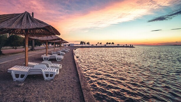 Ein Strand mit Liegestühlen und einem Reetdachschirm, ruhiges Meer und ein Sonnenuntergangshimmel.