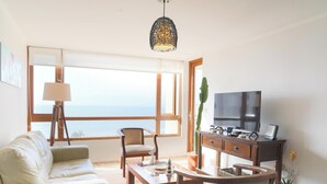 A living room with a sofa, a chair, a coffee table, a TV, and a large window with a view of the sea.