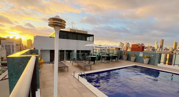 A rooftop pool with a city skyline in the background, surrounded by outdoor seating and a modern building. - Magnifique - Miramar Hospedagens  (João Pessoa)