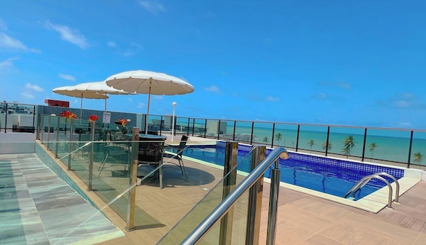 A rooftop pool area with glass railings, a pool, and a view of the ocean. - Magnifique - Miramar Hospedagens  (João Pessoa)