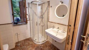 Een badkamer met een douchecabine van glas, een wastafel, een ronde spiegel en een raam met geopende luiken.