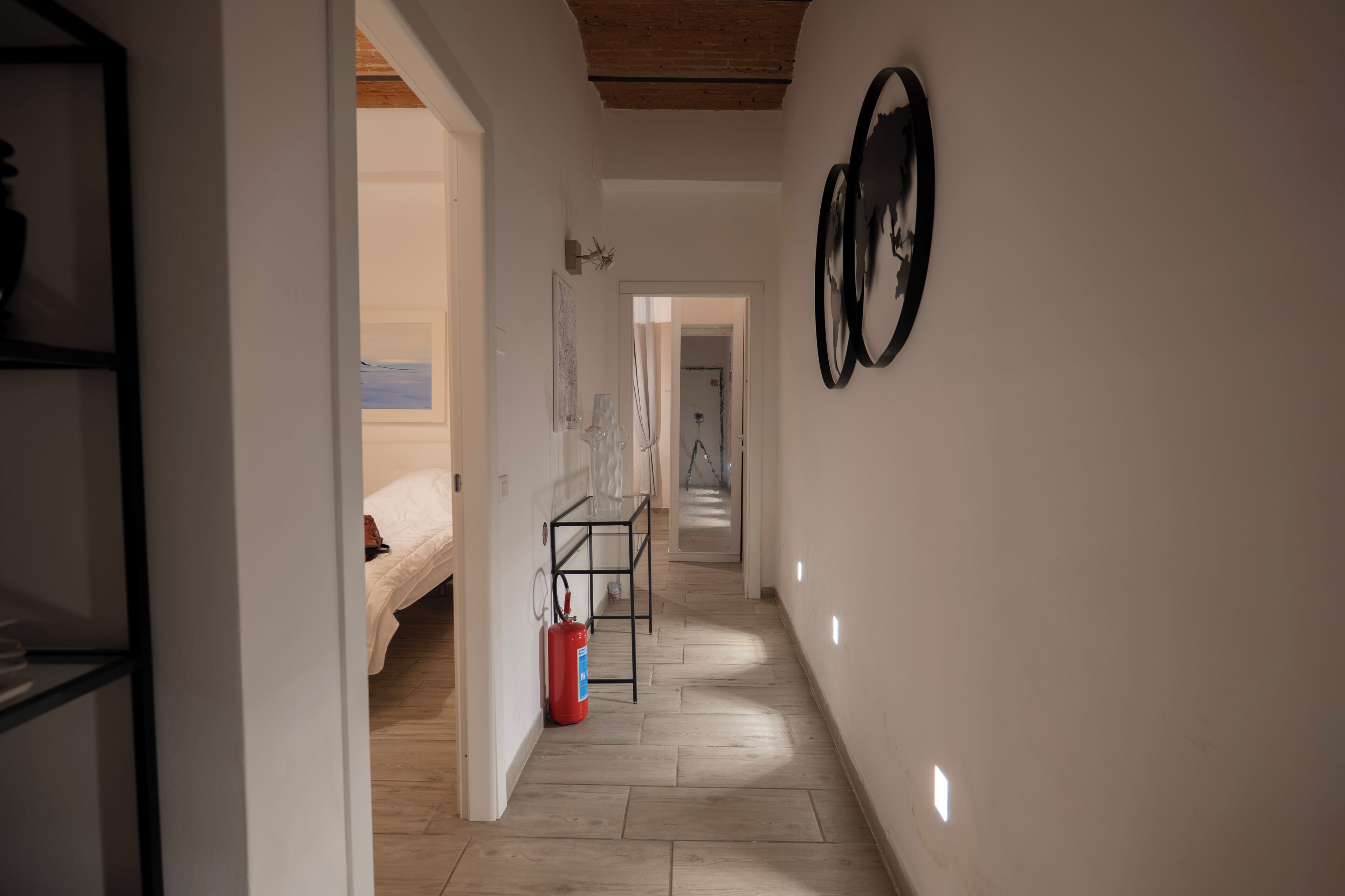 Un couloir avec un lit, une table sur laquelle se trouve un extincteur, et un mur orné de deux décorations circulaires.